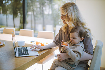 madre con niño de un año comiendo una mandarina en el regazo y trabajando en el portátil